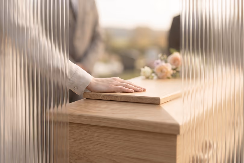 Ruka spočívá na dřevěné rakvi s květinami v rozostřeném pozadí.