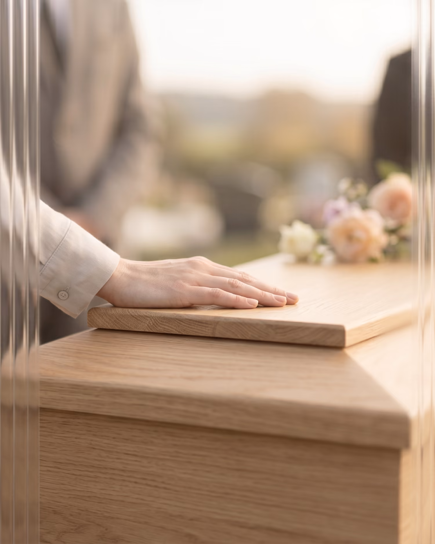 Ruka spočívající na dřevěné rakvi s rozmazanými květinami v pozadí.