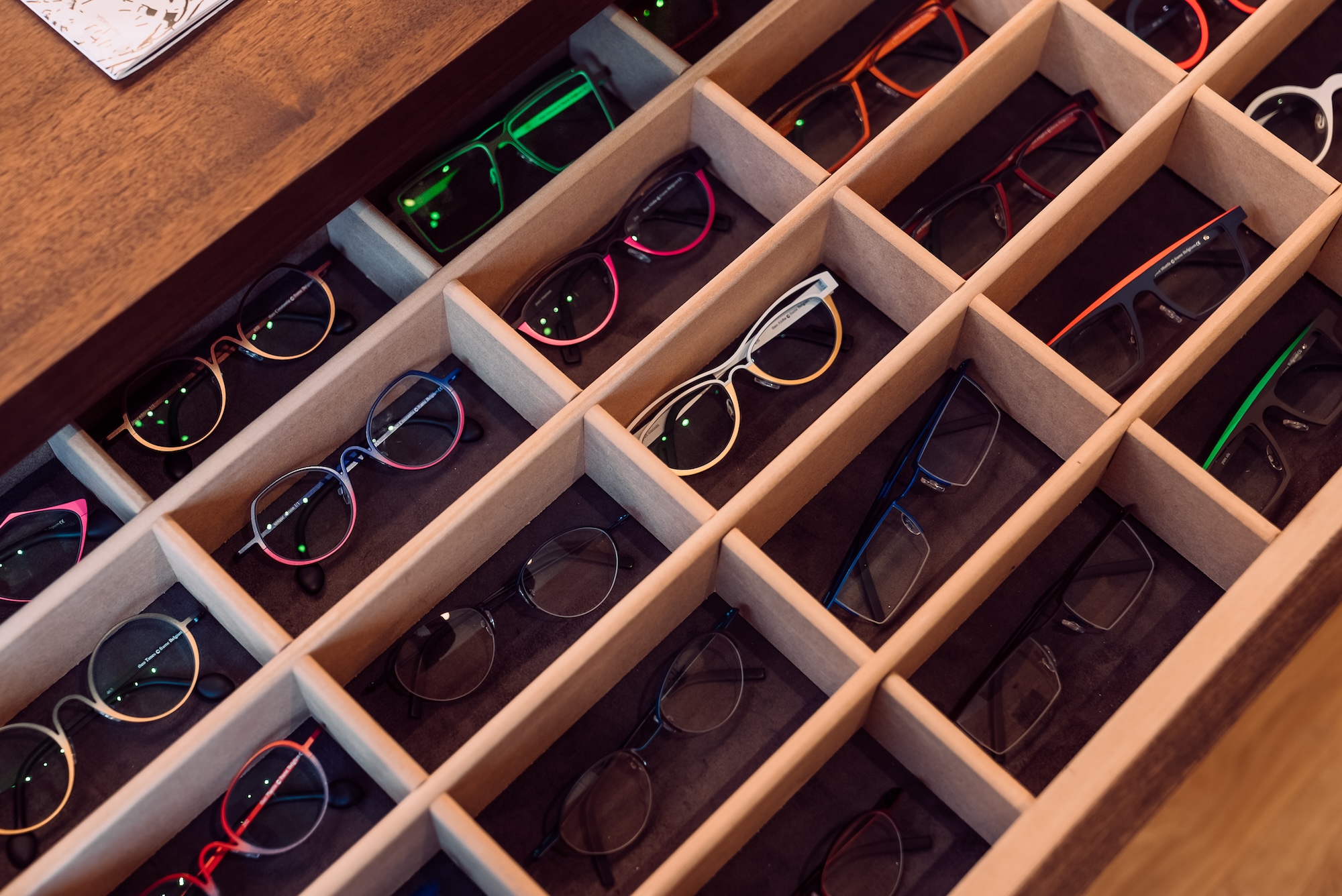 Multiple eyeglass frames of various colors neatly arranged in a wooden display case with compartments.