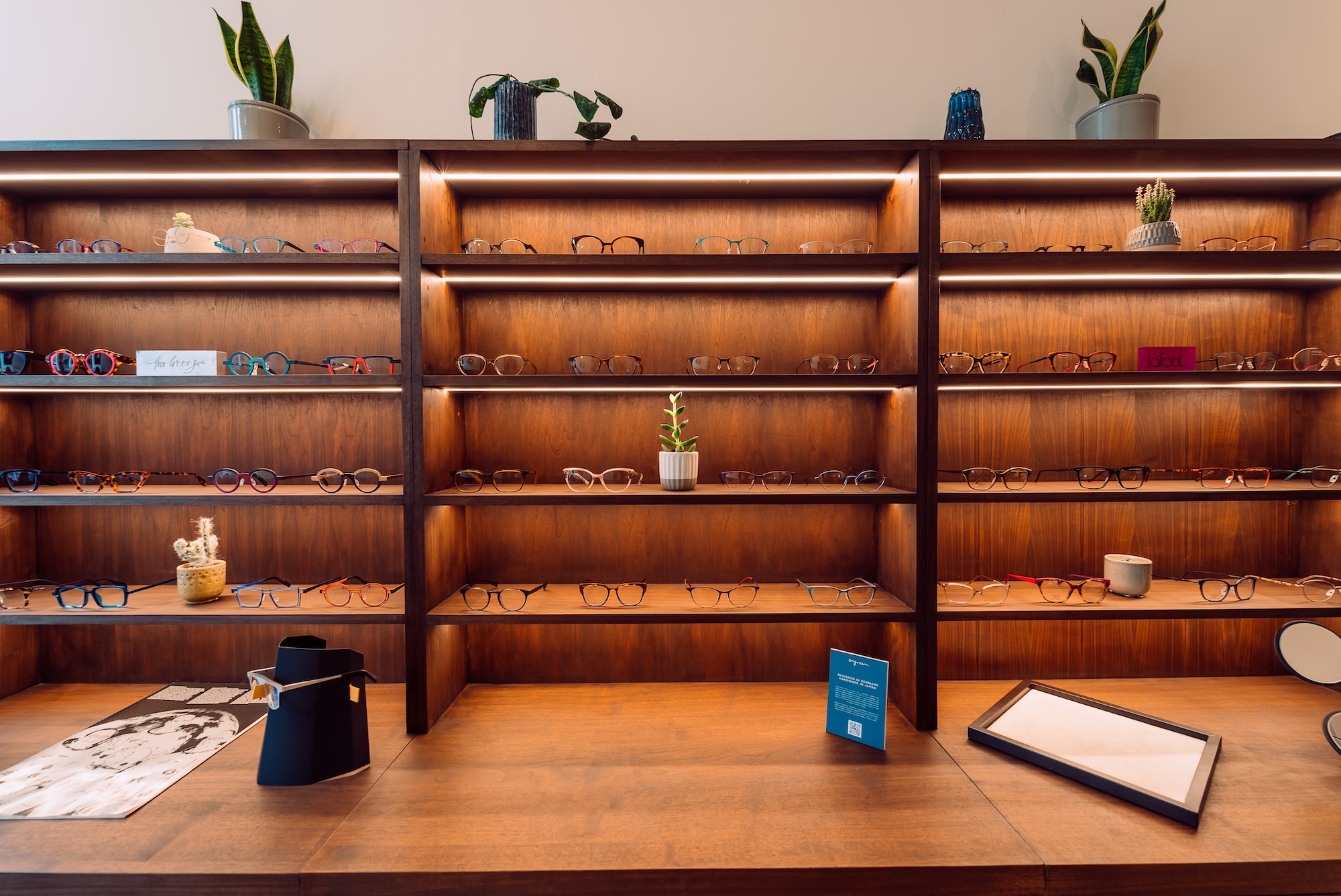 Wooden shelves with a variety of eyeglasses and small potted plants in a well-lit optical store.