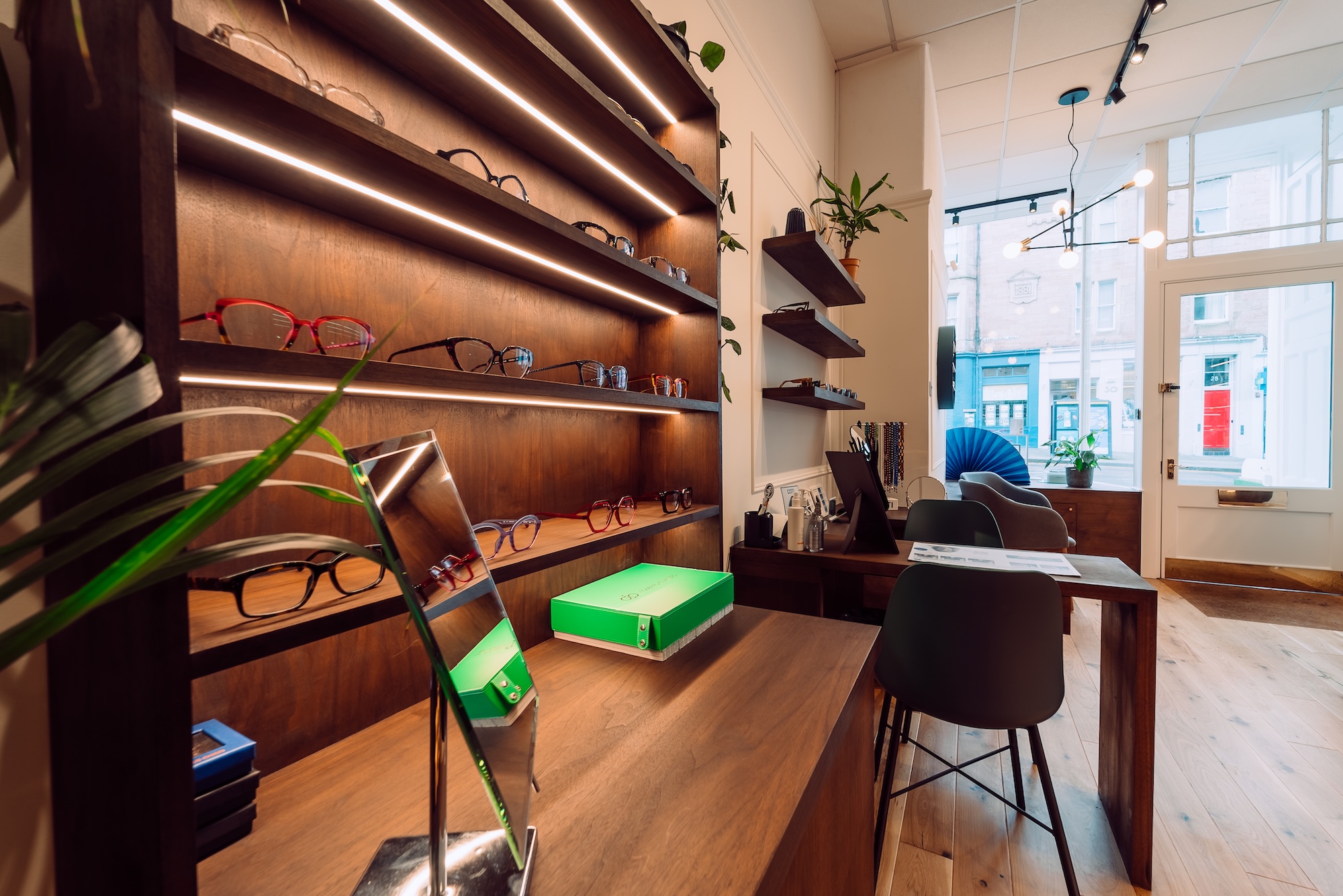 Interior of an eyewear store showing wooden shelves with glasses, a mirror, and seating area by the window.
