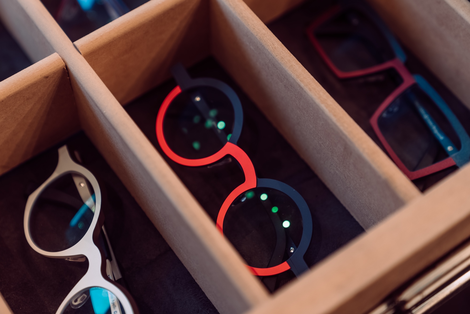 Three pairs of eyeglasses in a cardboard divider, including round black-red and rectangular red frames.