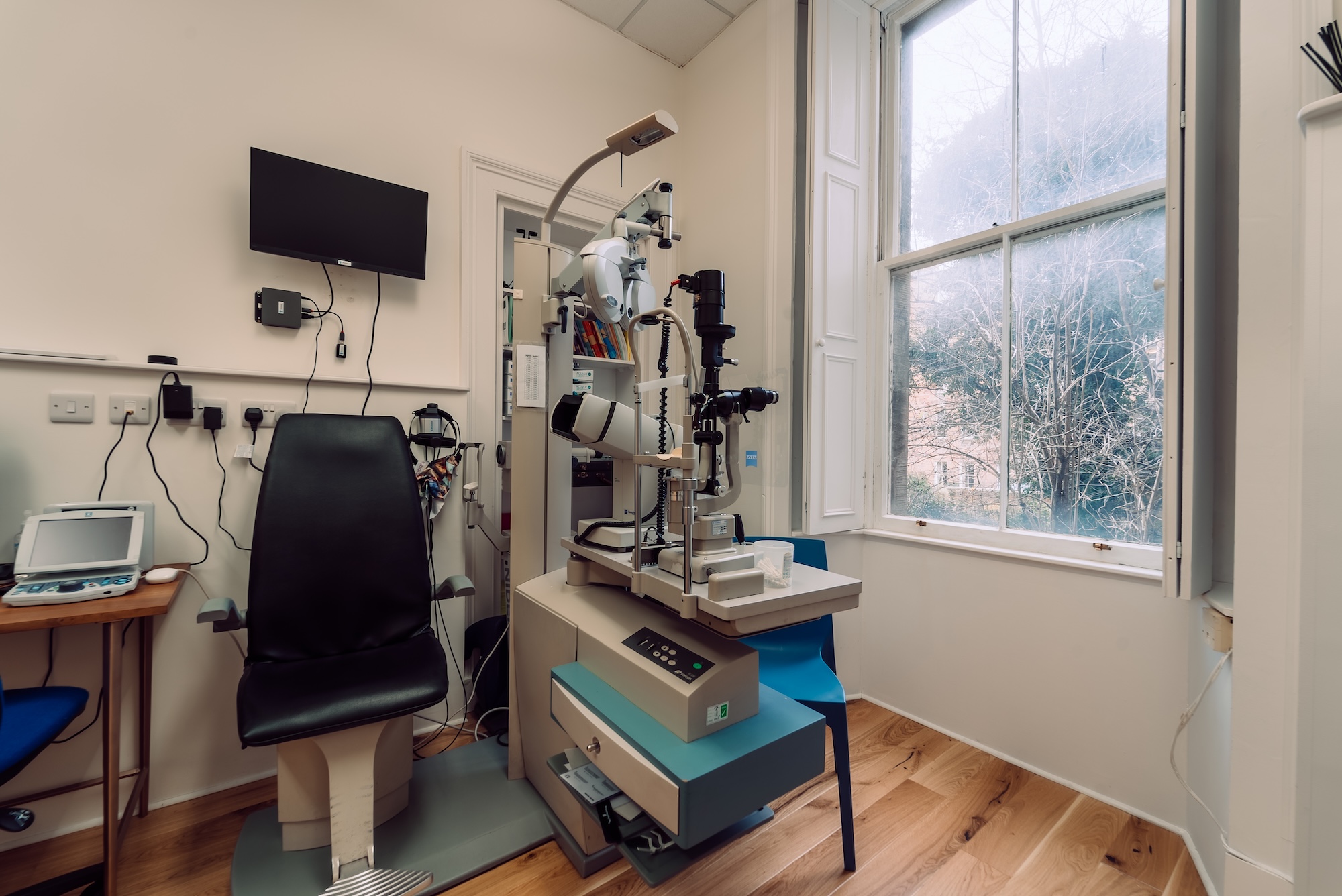 Optometrist exam room with an eye test chair, eye exam machine, and a large window showing trees outside.