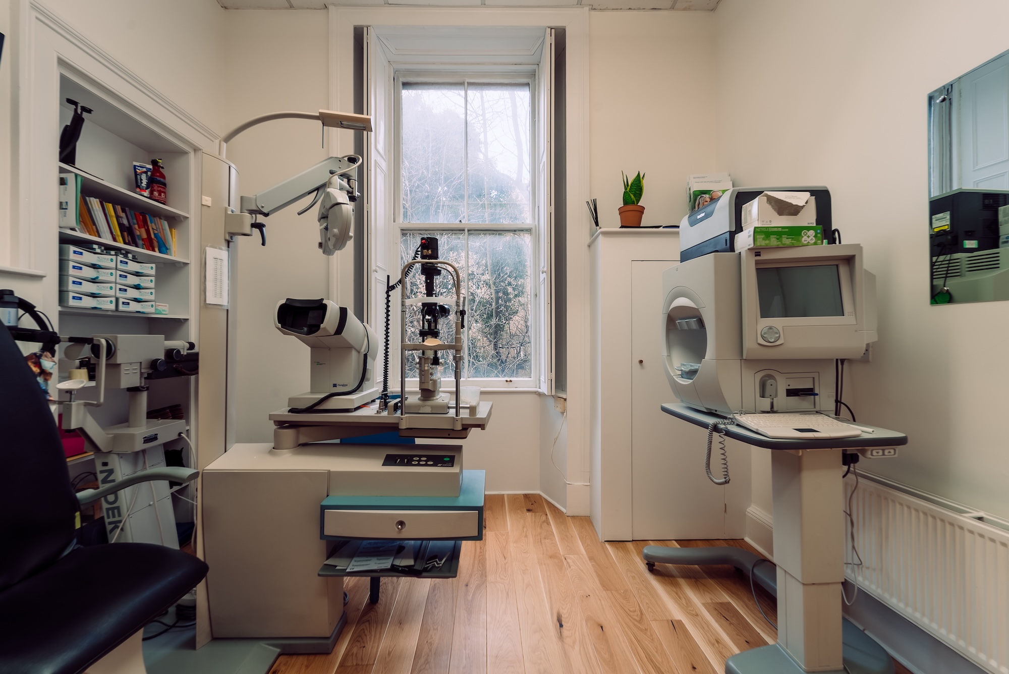 Eye exam room with optical machines, chair, wooden floor, a window, and a plant on a cabinet.