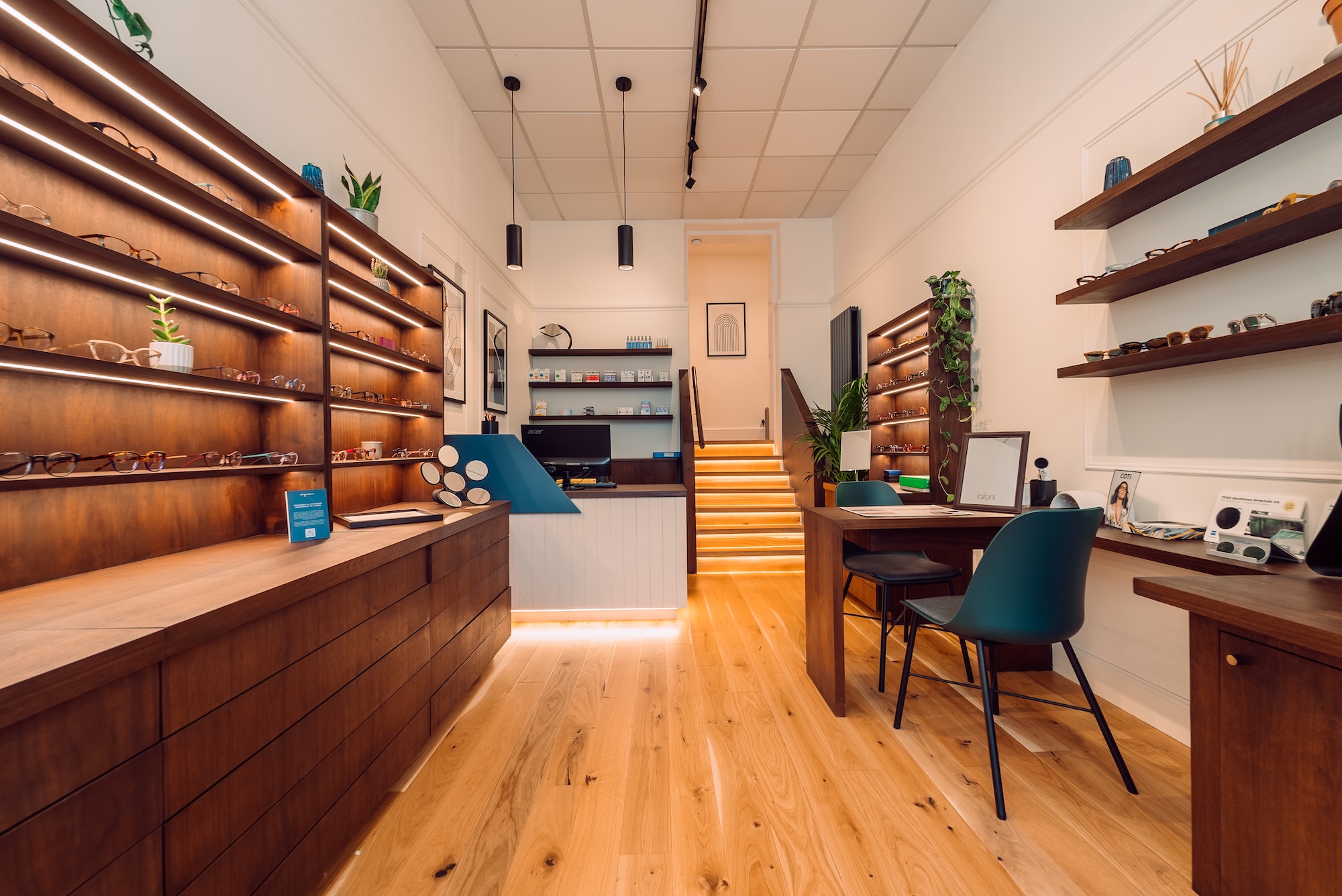 Modern optician store interior with wooden shelves displaying eyeglasses and a consultation desk with chairs.