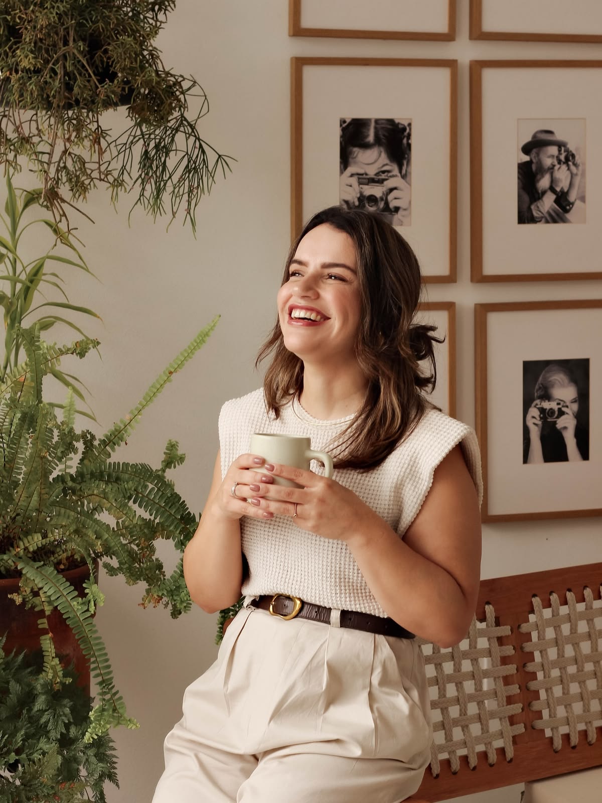 Smiling woman holding a mug, seated near plants and framed black-and-white photos on a wall.