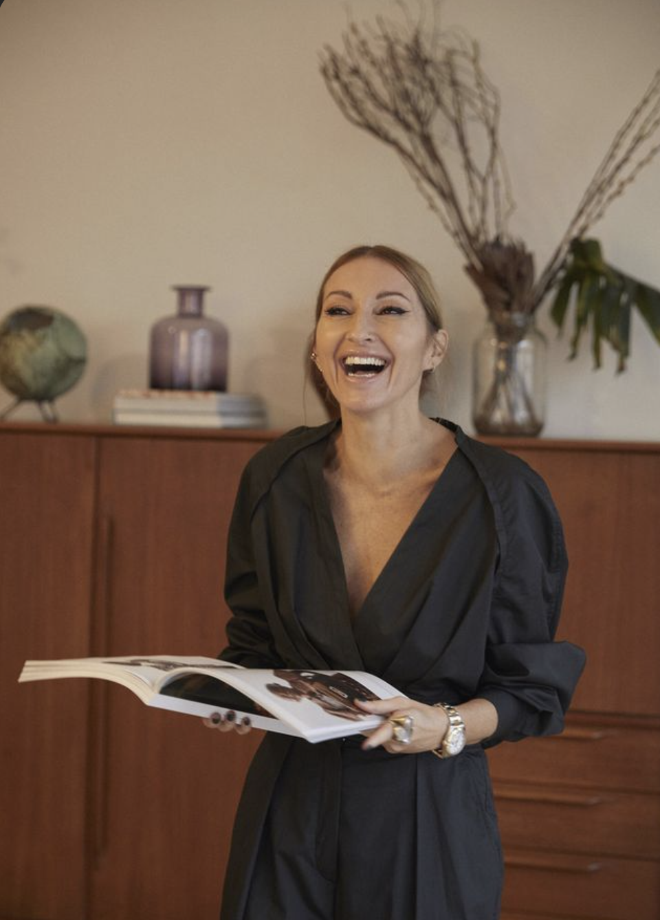 Smiling woman in black holding an open book with home decor in the background.