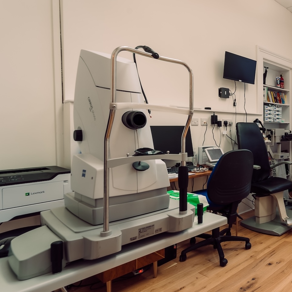 Optometry diagnostic equipment on a table with chairs and monitors in an eye exam room.