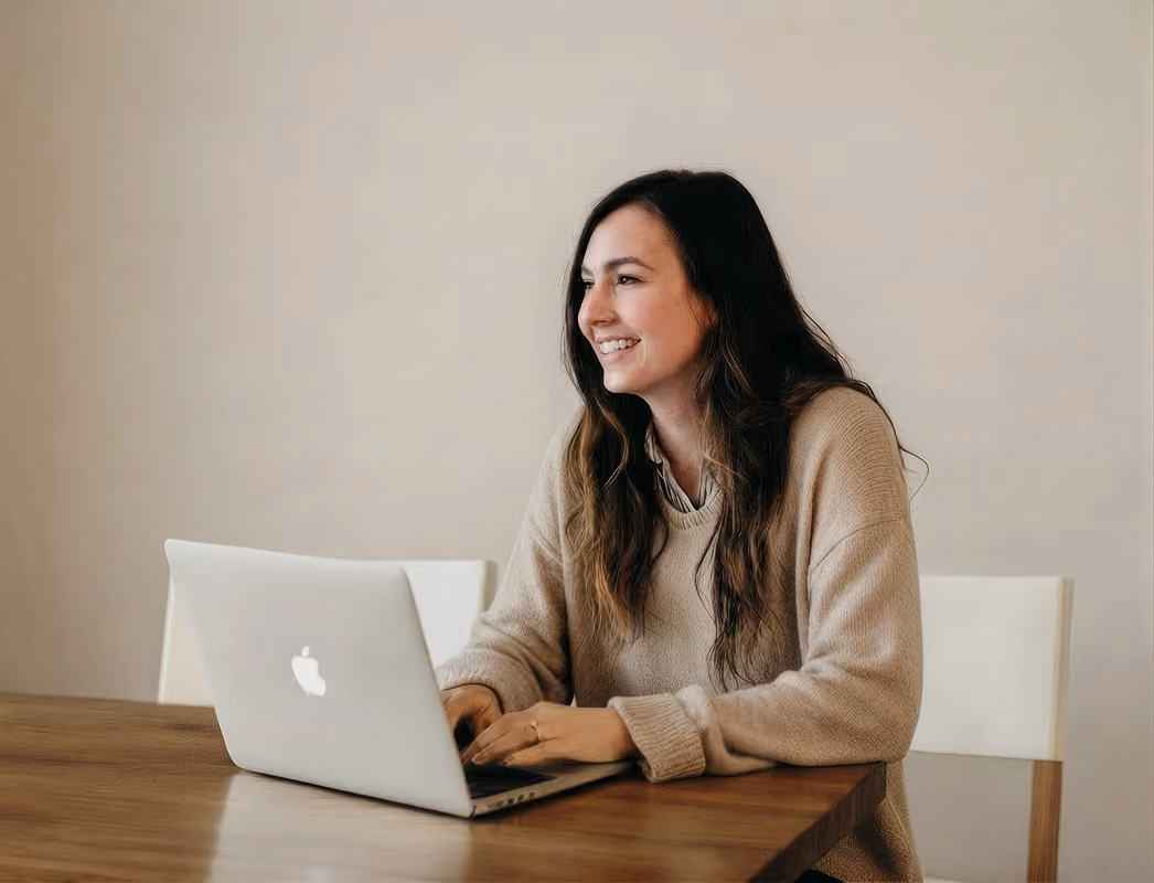 Smiling woman with long hair using a MacBook laptop at a wooden table against a beige wall.
