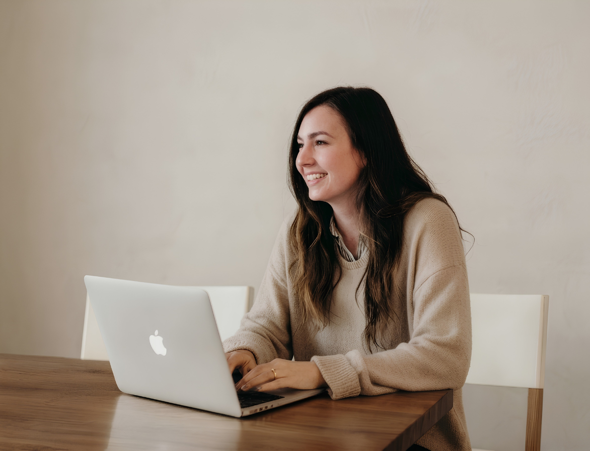 Smiling woman with long hair using a MacBook laptop at a wooden table against a beige wall.