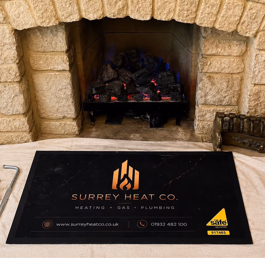 Black Surrey Heat Co. safety mat with logo by a stone fireplace with glowing gas logs.