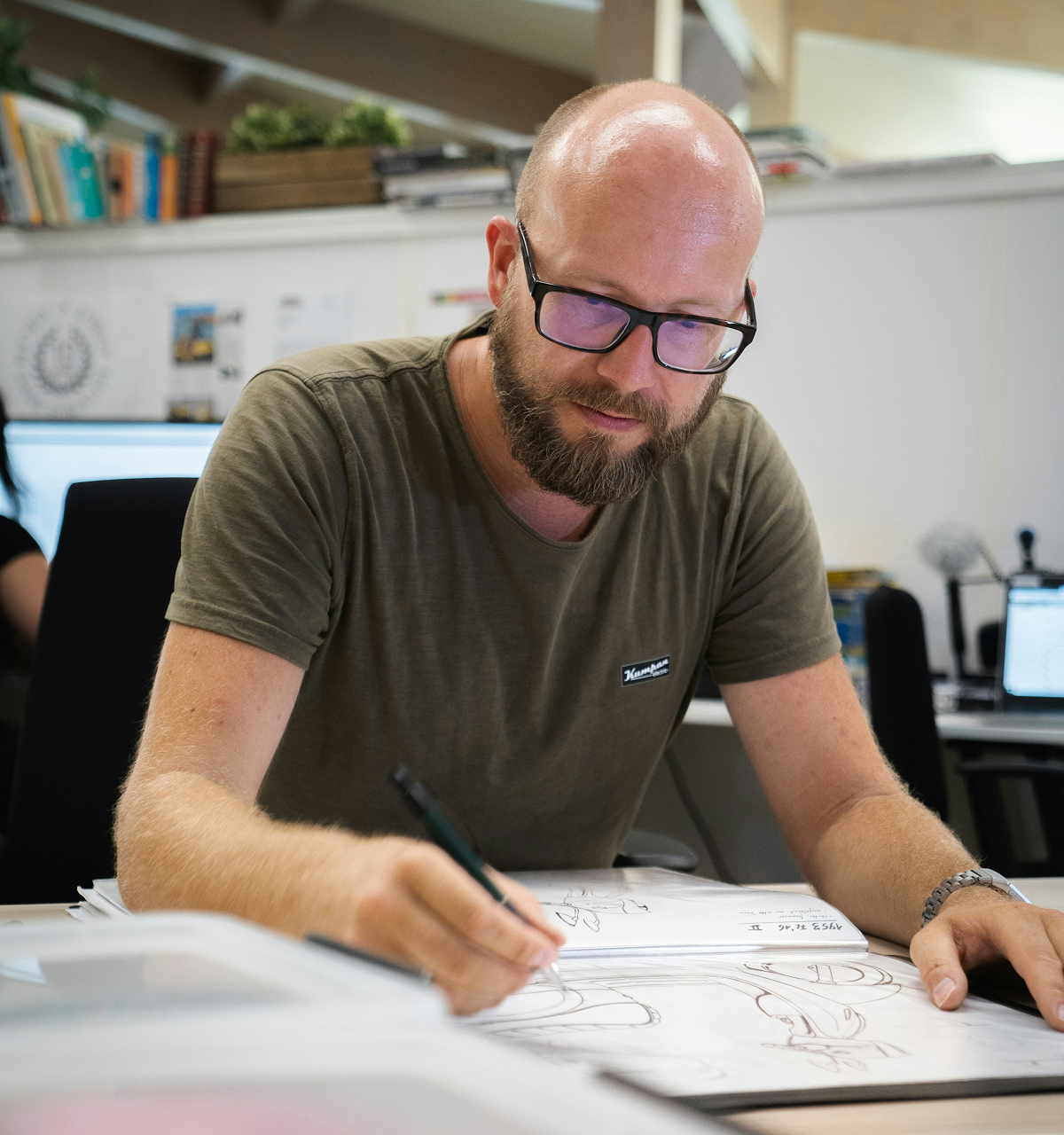 Bearded man wearing glasses drawing sketches on paper at a desk in an office.