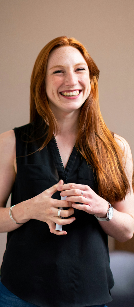 Smiling red-haired woman wearing a black sleeveless blouse, holding an object in her hands.