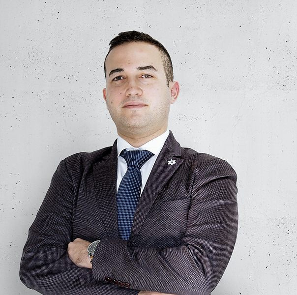 Man in a suit and tie with arms crossed, standing confidently against a light gray wall.
