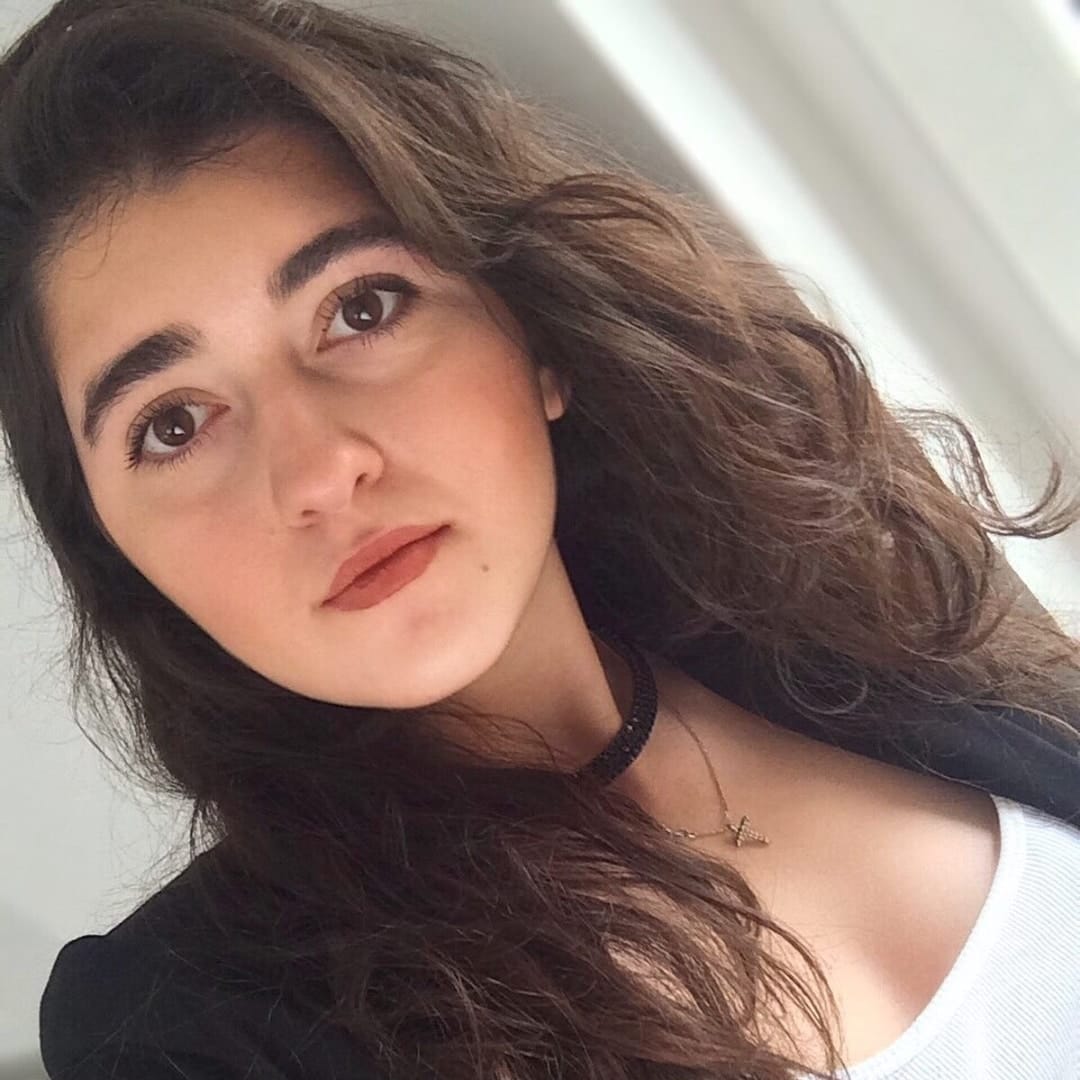Close-up of a woman with long wavy brown hair, dark eyes, and natural makeup wearing a black choker.