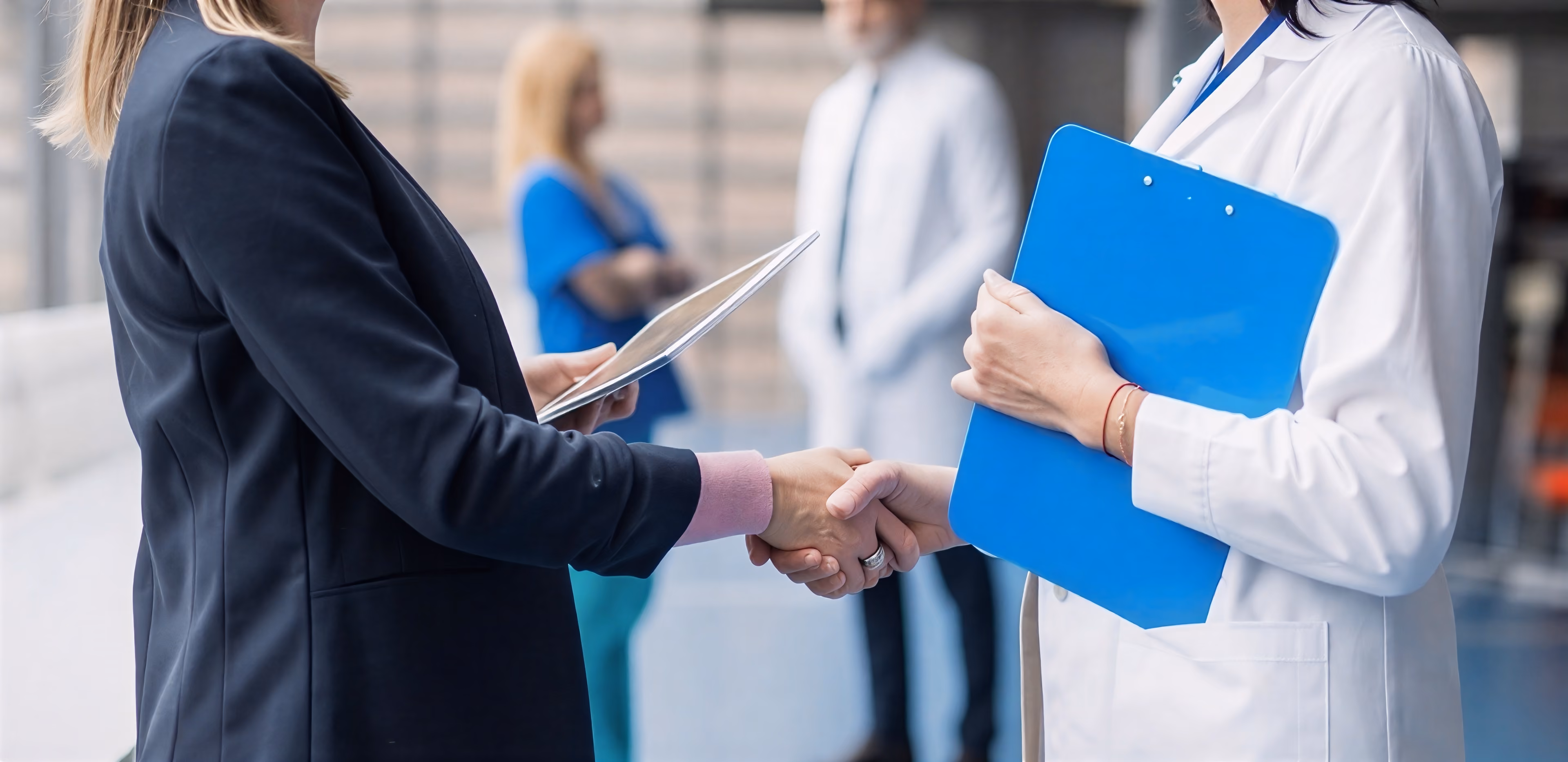 Businessperson shaking hands with a doctor holding a blue clipboard in a professional setting.