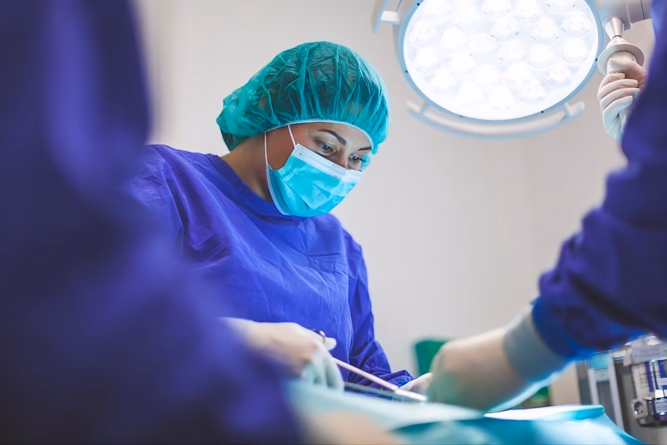 Healthcare professional in surgical scrubs and mask focused on a procedure under bright operating room light.