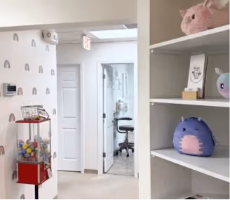 Interior of a room with a white door, chair, shelves with stuffed toys, and a red gumball machine.