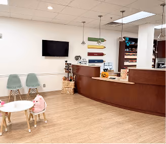 Reception area with curved wooden desk, small table with toys, two chairs, and colorful directional signs on wall.