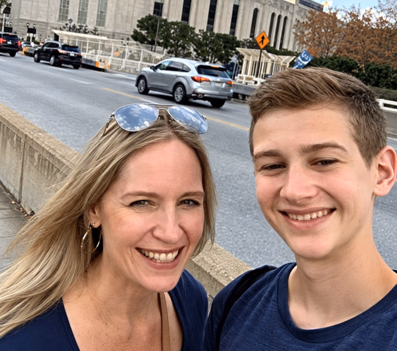 Smiling woman and teenage boy taking a selfie outdoors near a street with cars in the background.