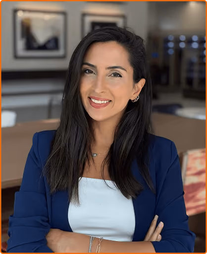 A smiling woman with long dark hair wearing a navy blazer and white top in a bright room.