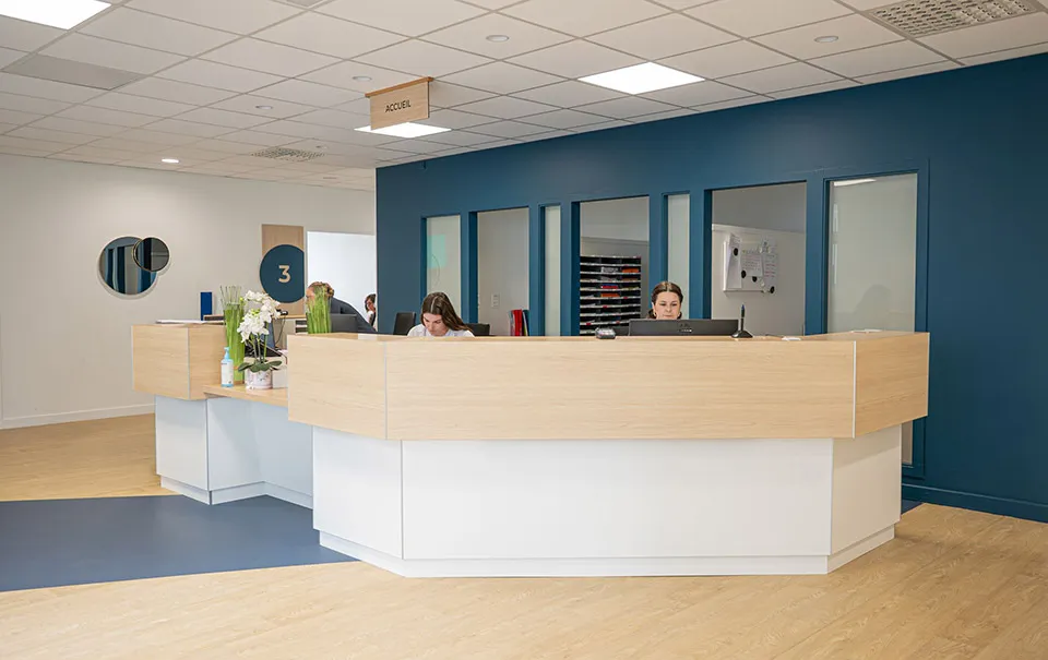 Modern reception area with a wooden desk and two receptionists working on computers, a dark blue wall behind with windows and a hanging sign labeled ACCUEIL, light wooden floor with blue carpet section, and decorative plants on the desk.