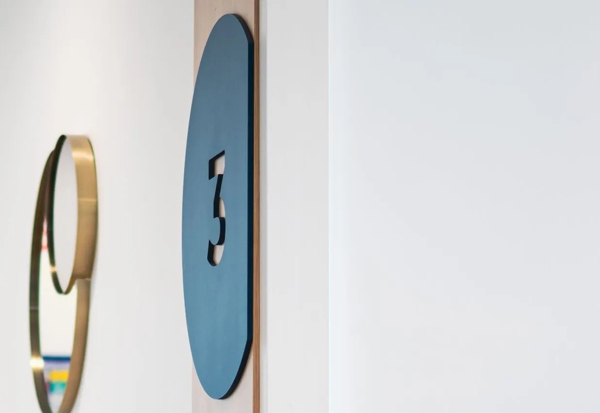 Blue oval room number sign with the number 3 mounted on a light-colored wall, next to a circular gold-framed mirror.