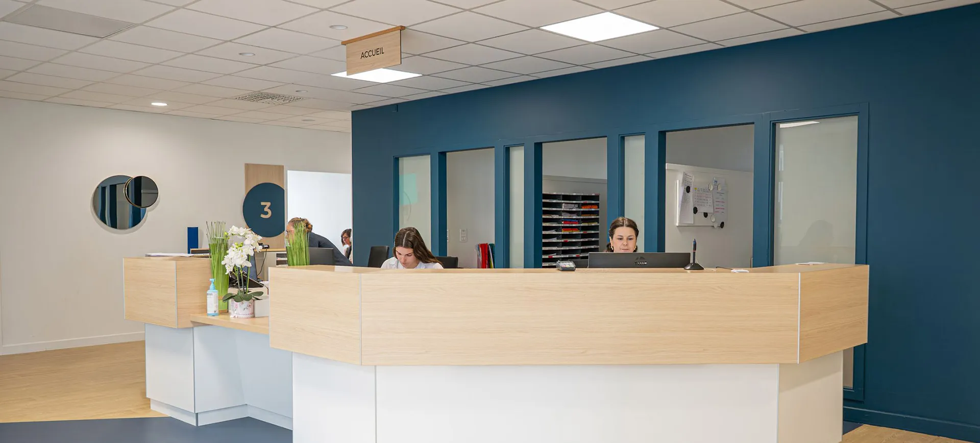 Modern reception area with light wooden desk and blue accent wall, two receptionists working at computers behind the desk, and a sign hanging from the ceiling labeled 'ACCUEIL'.