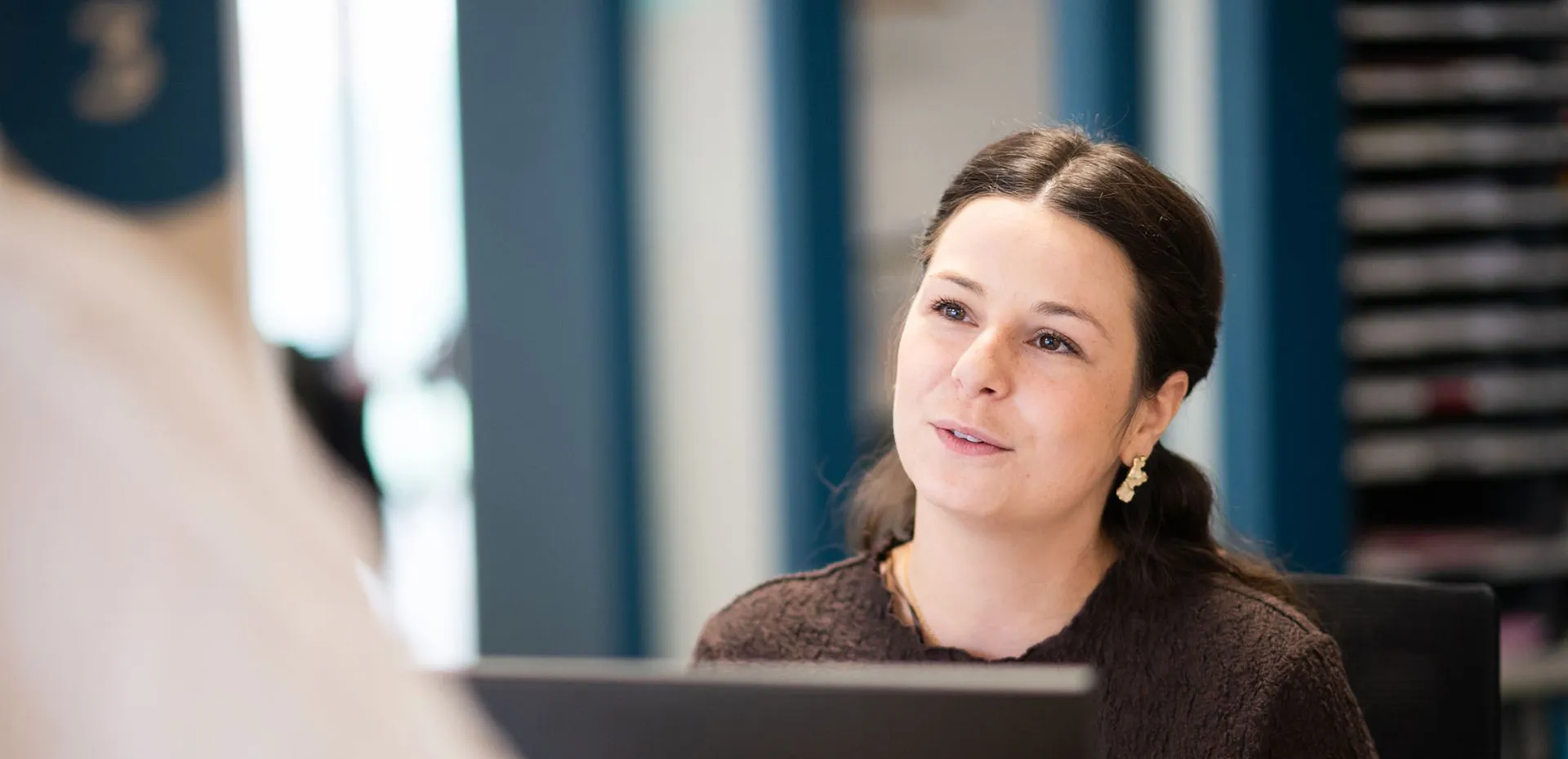 A woman with dark hair pulled back, wearing gold earrings and a dark textured top, looks attentively at someone in an indoor office setting.