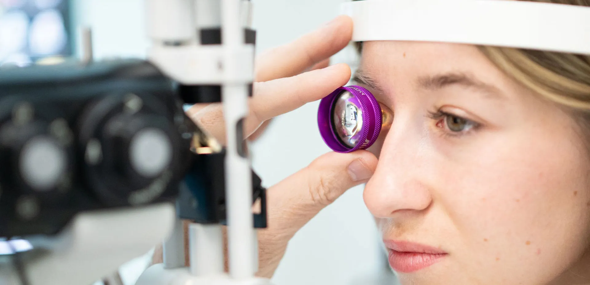 A woman undergoing an eye examination using a lens held by a hand in front of her eye, with an eye exam device visible next to her face.