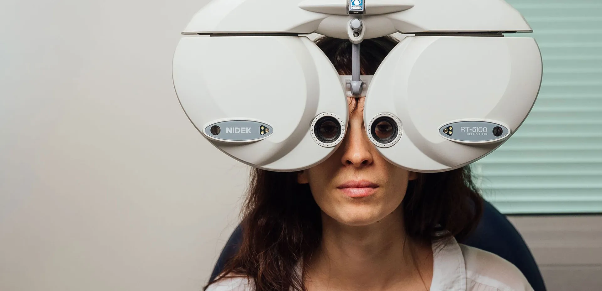 Woman undergoing an eye exam using a Nidek RT-5100 refractor device, with her eyes visible through the machine's lenses.