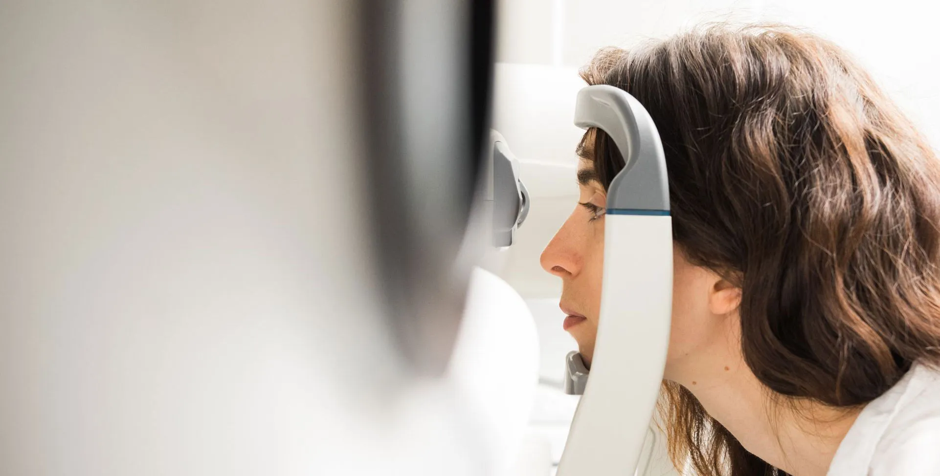 Woman with brown hair undergoing an eye examination using a medical device, facing right with her chin resting on the instrument.
