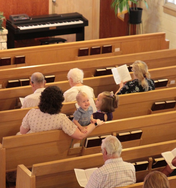 A little girl at Trinity Lutheran Church in Paola, Kansas turns around in a light colored pew to smile at camera that is coming from above. She is surrounded by other parishioners who are worshiping.