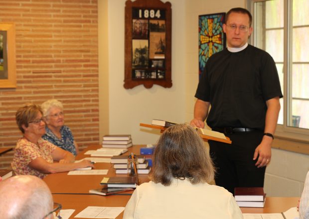 Pastor Joshua Woelmer leads his members at Trinity Lutheran Church in Paola in a study through the bible. They are in a fellowship hall with art on the walls, and Pastor Woelmer is wearing a short sleeved black clerical shirt with a collar. 