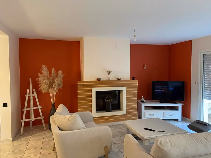 Salon avec deux fauteuils beige, cheminée encadrée de bois, TV sur meuble blanc et murs rouges.