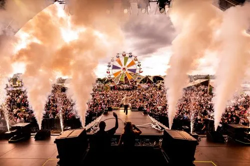 Een DJ en een man op een podium voor een massa mensen met rook- en vuurwerkshows en een kleurrijk reuzenrad aan de horizon.