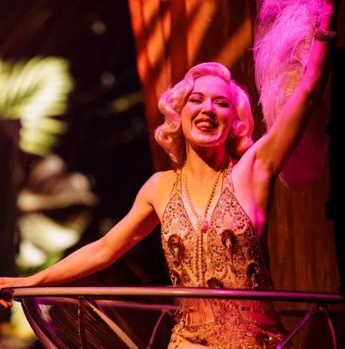 Vrouw in glamourachtige vintage avondjurk met parelketting lacht terwijl ze haar arm omhoog houdt in theatrale setting.