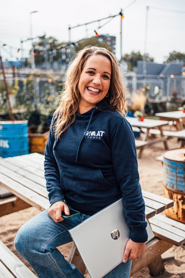 Vrouw met een donkerblauwe hoodie en spijkerbroek lacht terwijl ze op een houten picknicktafel zit en een zilveren laptop vasthoudt in een buitenruimte.