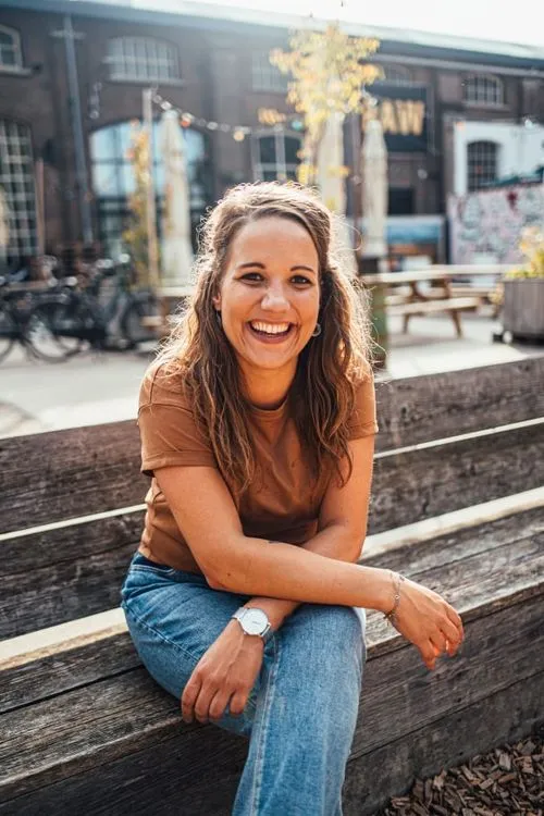 Vrolijke vrouw met lang haar, bruin shirt en blauwe jeans, zittend op een houten bankje buiten in stedelijke omgeving.