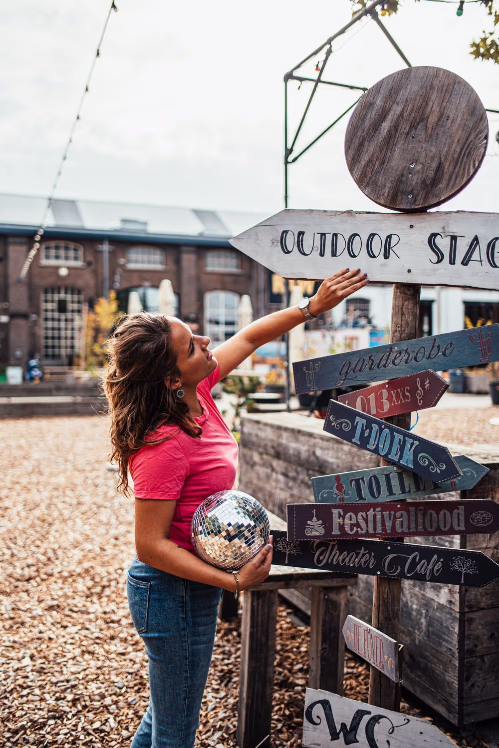 Vrouw met een discobal die een pijlbord met diverse festivalrichtingen bekijkt, waaronder 'Outdoor Stage' en 'Festivalfood'.