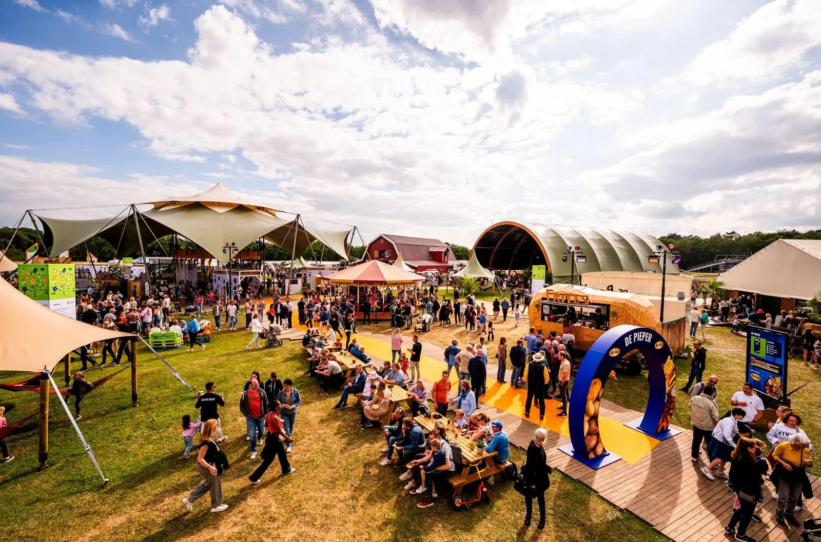 Druk festivalterrein met mensen, diverse tenten, eetkraampjes en zitplaatsen op gras tijdens een zonnige dag.