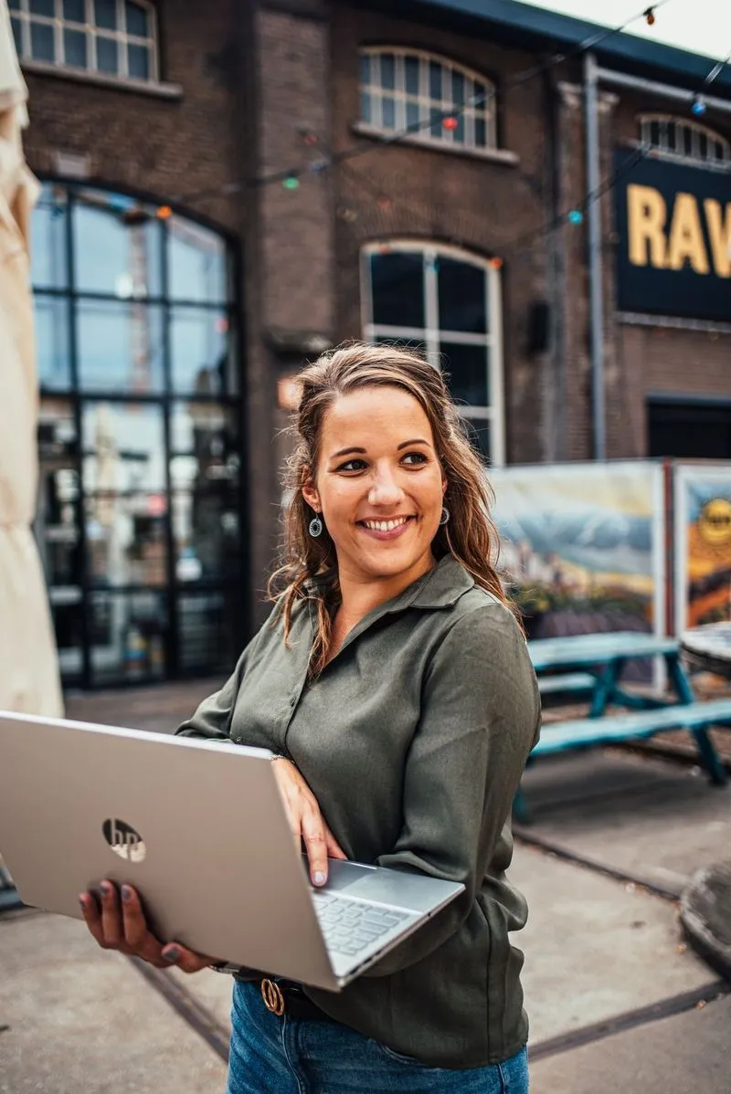 Vrouw met donkere blouse lacht terwijl ze een zilverkleurige HP-laptop vasthoudt in een stedelijke buitenomgeving.