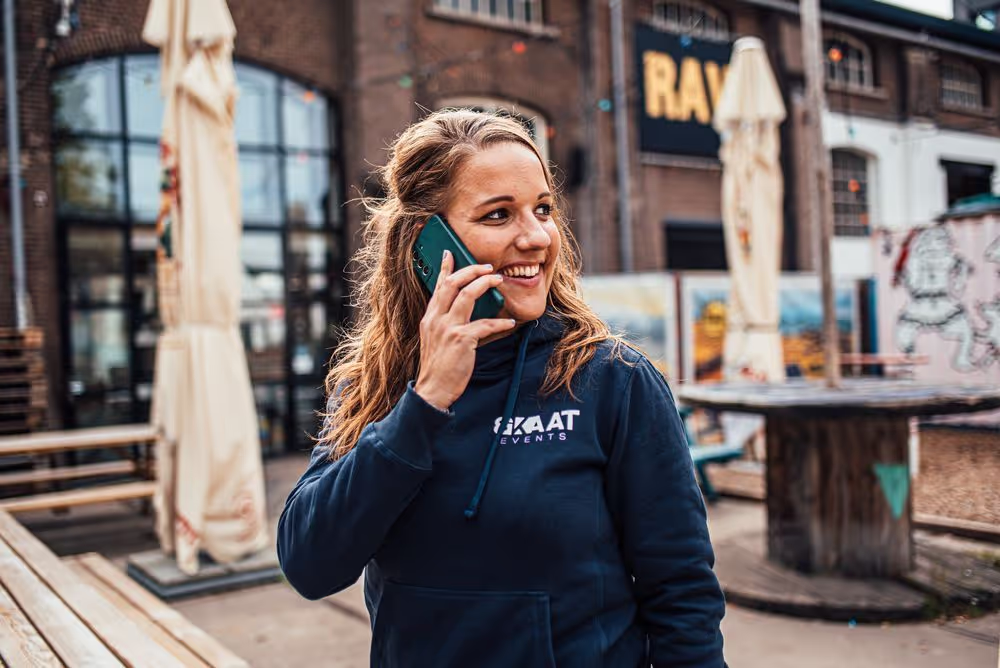 Vrouw in een donkerblauwe hoodie met tekst 'KAAT EVENTS' die glimlacht terwijl ze buiten op een mobiel telefoontje spreekt.