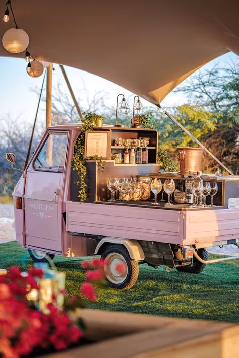 Roze driewielige bestelwagen omgebouwd tot een mobiele bar met glazen, flessen en decoratie onder een witte luifel buiten.