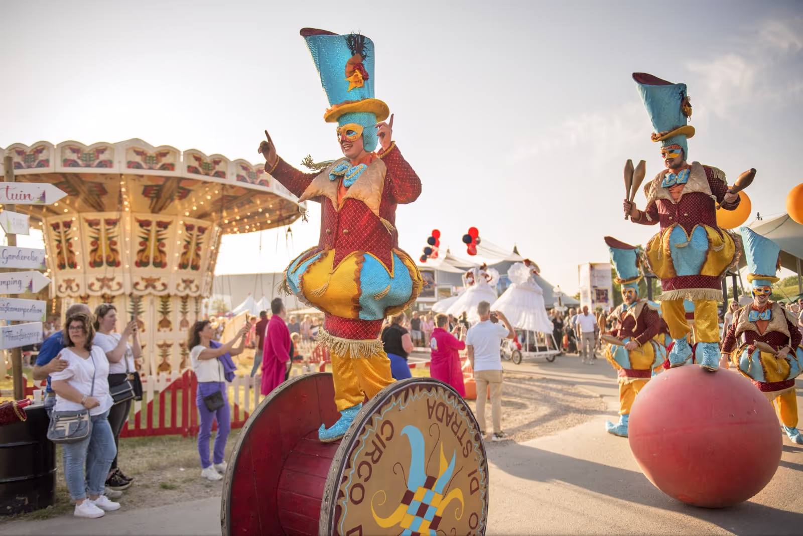 Performers in colorful jester costumes, including tall blue hats and yellow-blue outfits, balancing on large props and balls at an outdoor festival with spectators and carousel in the background.