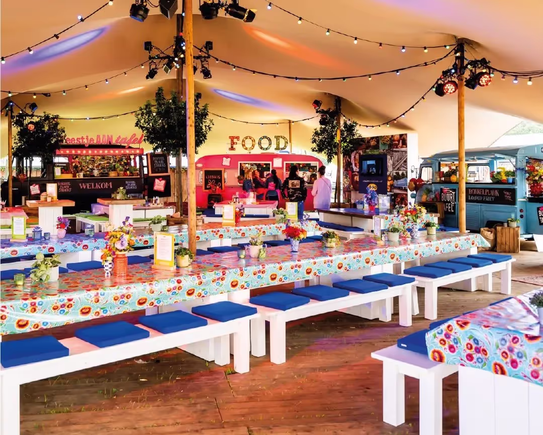 Grote feesttent met lange tafels bedekt met kleurrijke tafelkleden, blauwe kussens op banken, bloemen en eetkraampjes met verlichting.