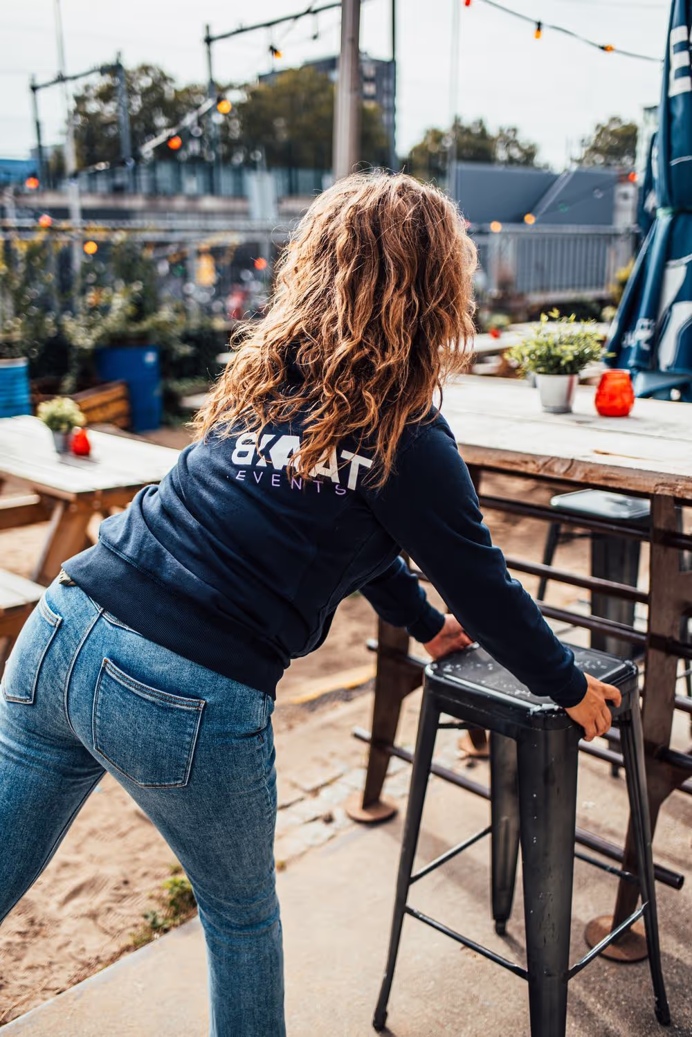 Vrouw in een blauwe trui met opdruk '8KAT EVENTS' die buiten een zwarte barkruk verplaatst.
