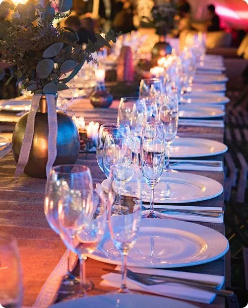 Elegante gedekte tafel met witte borden, glazen en een vaas met bloemen, sfeervol verlicht voor een evenement.