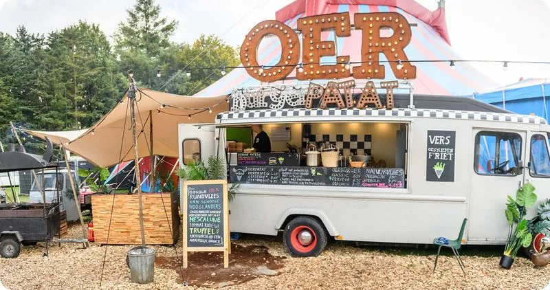 Witte foodtruck met grote OER patat-verlichting en menuborden, omgeven door tenten en bomen.