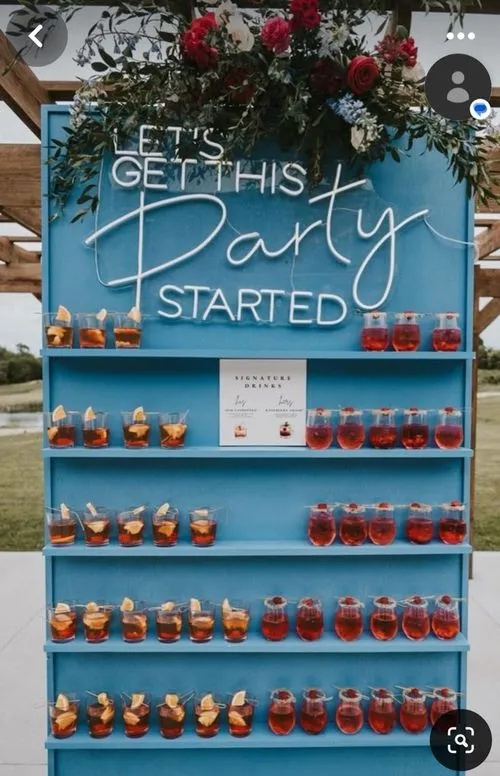 Blauwe dranktafel met meerdere glazen met gele en rode cocktails, bovenaan versierd met bloemen en een neonbord met de tekst 'Let's Get This Party Started'.